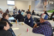 Students sit around a table talking with a Native American mural in the background.