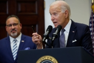 President Biden stands at a podium. A blurred-out Miguel Cardona is in the background.