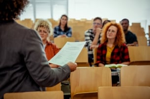 Close-up of a teacher's hand holding a paper while they talk to a group of college students in a lecture-style hall