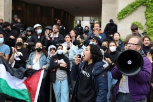  A group of twenty or so protesters gathered in a doorway. In the left bottom corner of the frame some protesters hold an unfurled Palestinian flag. One speaker in front carries a bullhorn, while another appears to be shouting into a microphone. Police officers wearing helmets and face shields are standing in the background.