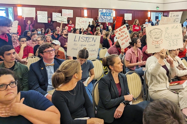 People sit in a large, full room, with some people standing in the back. Multiple individuals are holding protest signs, including one saying “We Demand Freeze Academic Transformation” and another saying “Gee You Drove Us in a Ditch.”